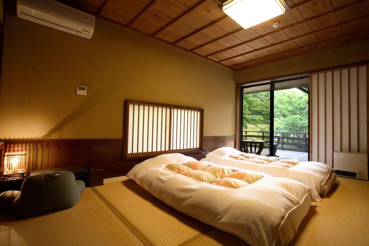 A traditional Japanese-style room with tatami flooring, a futon bed, a shoji screen, and a view of a forest through a sliding door.