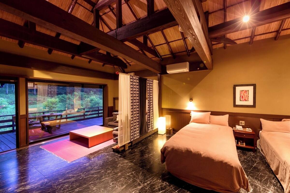 A hotel room with a large bed, a smaller bed, a desk, a chair, a window with a view of a deck, and a ceiling with wooden beams.