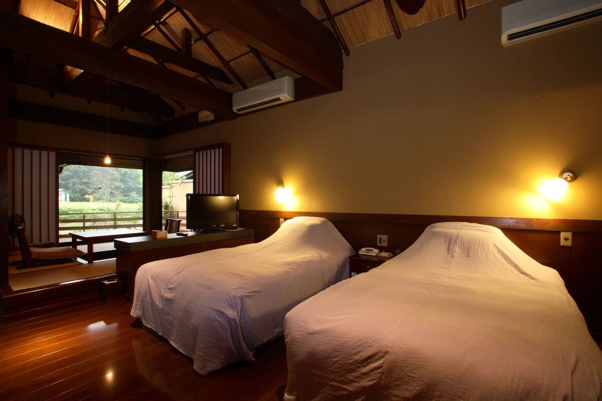 A hotel room with two beds, a wooden floor, a television, and a window with a view of greenery.