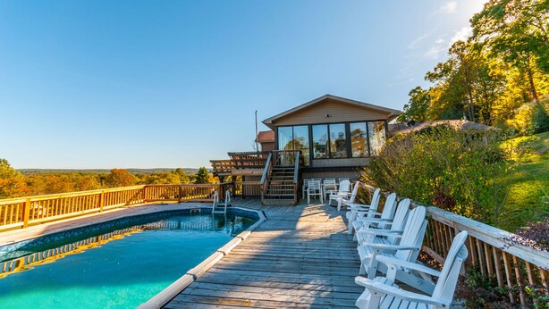 Una terraza de madera con piscina y sillas blancas, que conduce a un edificio con amplias ventanas y una terraza.