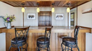 Una zona de bar con barra de madera, tres sillas negras, una jarra de metal con flores y dos cuadros con imágenes de animales en la pared.
