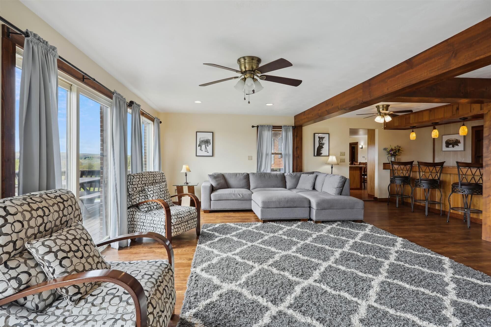 A spacious living area with a gray sectional sofa, patterned armchairs, and a patterned rug. There are ceiling fans, a dining area with bar stools, and a view of the outdoors through large windows.