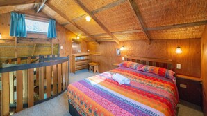 Une pièce chaleureuse avec un plafond en bois, un lit recouvert d’une couverture colorée, un bureau et un petit tabouret.