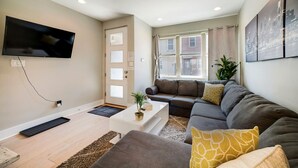 Ein modernes Wohnzimmer mit einem an der Wand montierten Flachbildfernseher, einer Sofagarnitur, einem Couchtisch und einem großen Fenster mit Vorhängen.