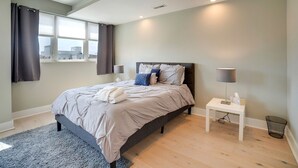 Ein modernes Schlafzimmer mit Bett, Nachttischen, einem Fenster mit Vorhängen und Blick auf Gebäude.
