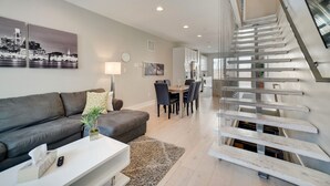 Ein modernes Wohnzimmer mit grauem Sofa, Couchtisch und Essbereich mit Holztisch und Stühlen. Eine Treppe mit Metallgeländer ist sichtbar.