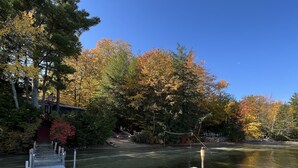 Eine Holzstege mit zwei Metallstühlen und einem Tisch, der zu einem ruhigen See führt, umgeben von Bäumen in Herbstfarben.