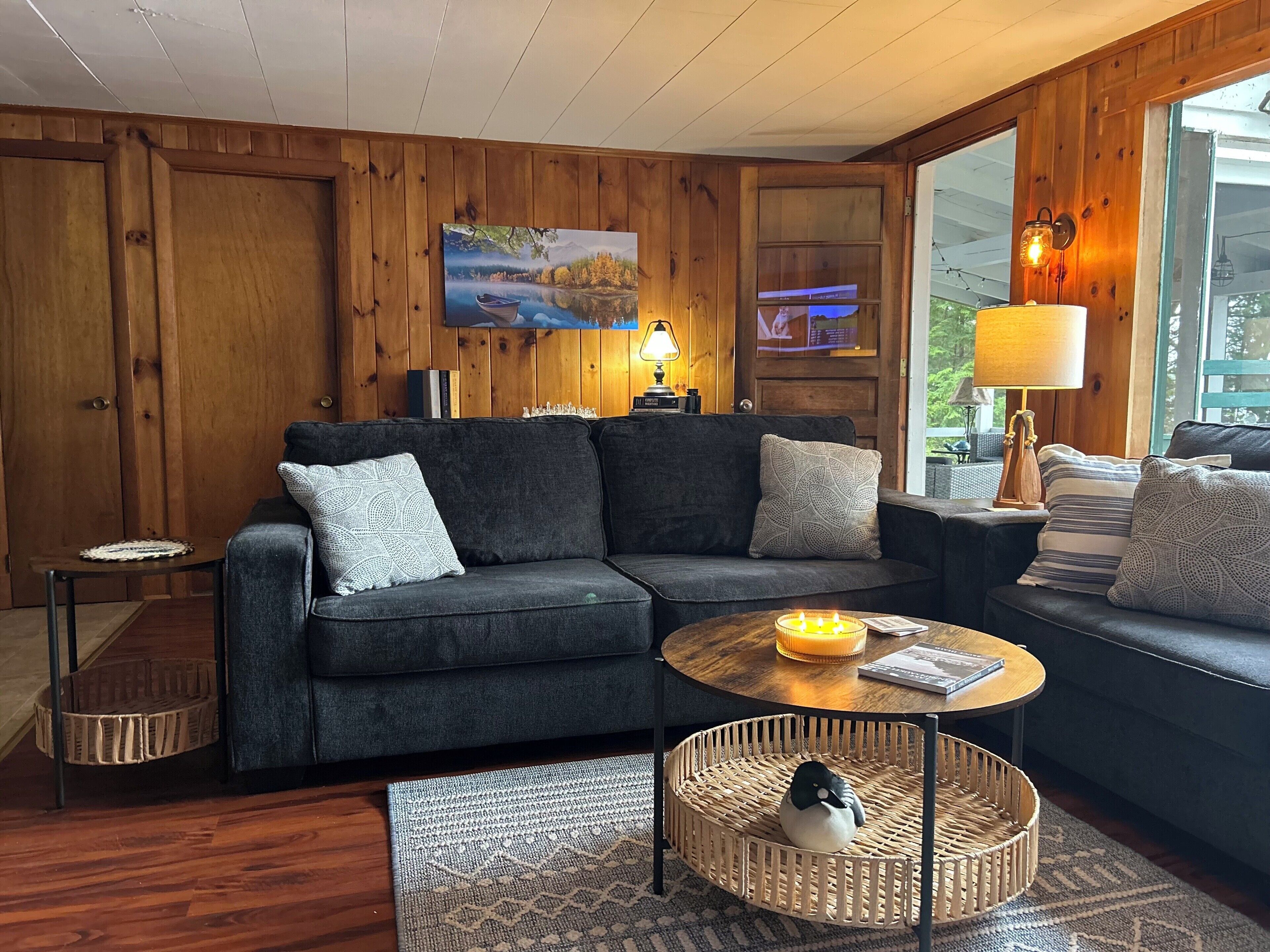 Ein gemütliches Wohnzimmer mit einem dunkelgrauen Ecksofa, einem runden Couchtisch mit Kerzen, einem gewebten Korb mit einem Plüschtier und einem großen Fenster mit Blick auf Grünflächen.