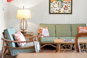 Et værelse med en grøn sofa, to træstole, et bord med en lampe og et blomsterbillede på væggen.
