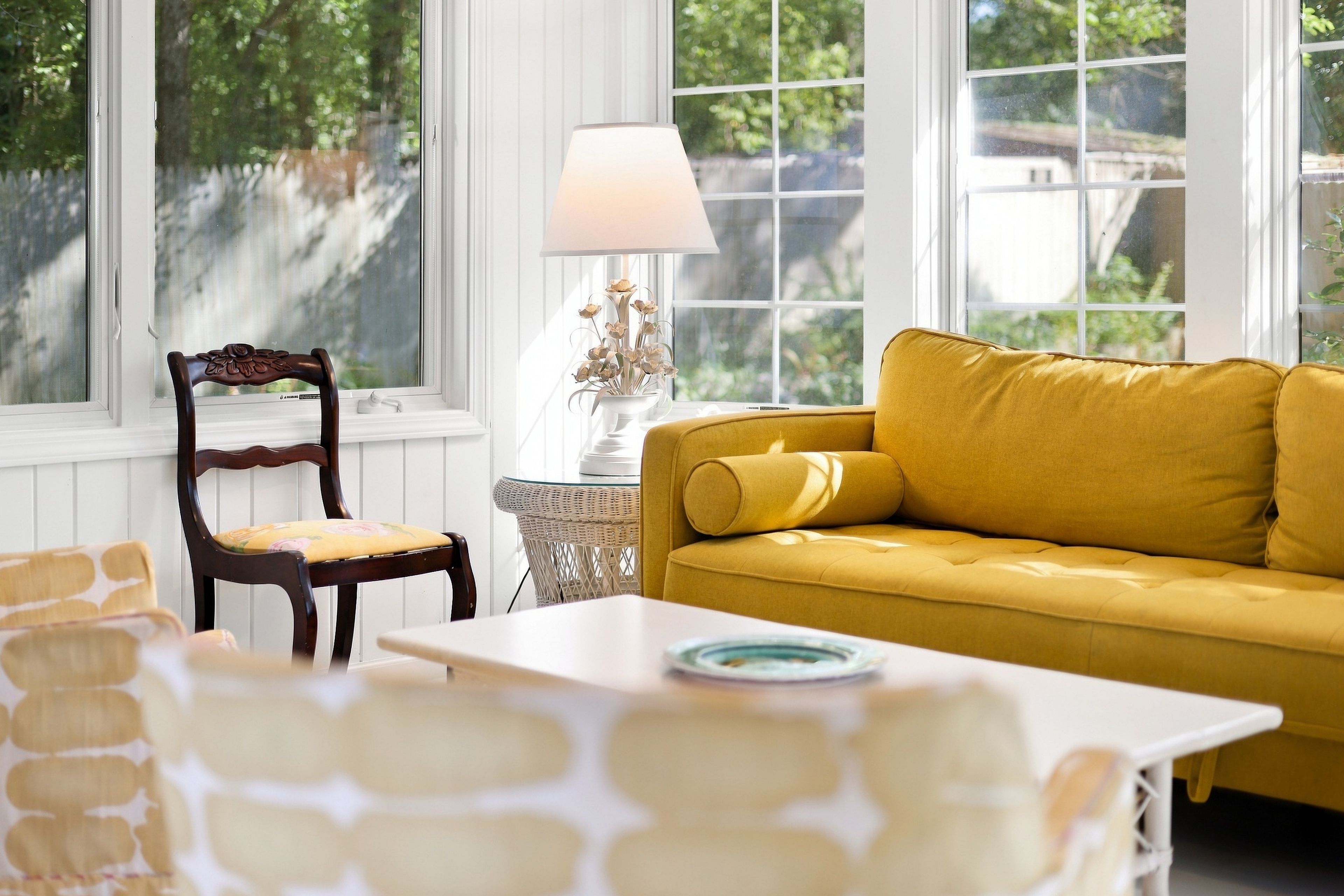 Una habitación con un sofá amarillo, una silla de madera con un cojín floral, una mesa blanca y una lámpara sobre una mesita auxiliar.