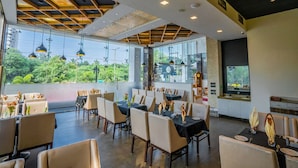 A modern restaurant with high ceilings, large windows, and a view of greenery. Tables are set with white tablecloths and candles.