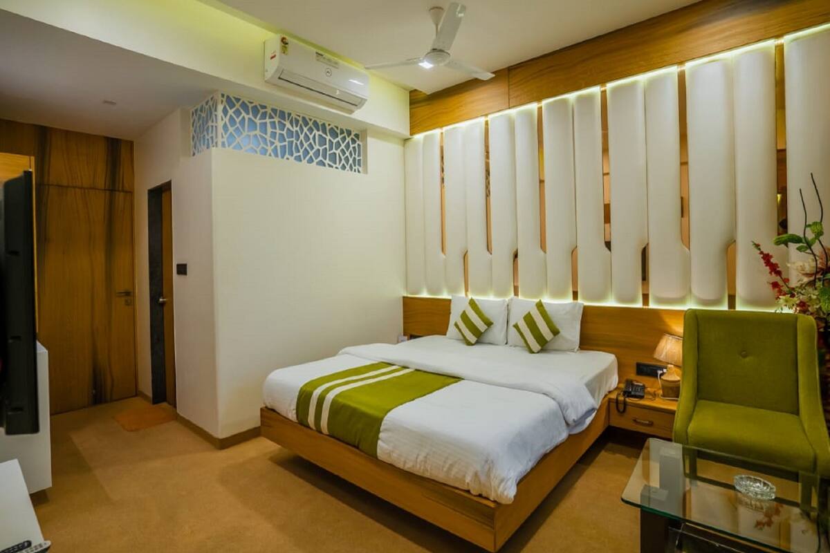 A modern hotel room with a bed, bedside table, lamp, green chair, and an air conditioning unit.
