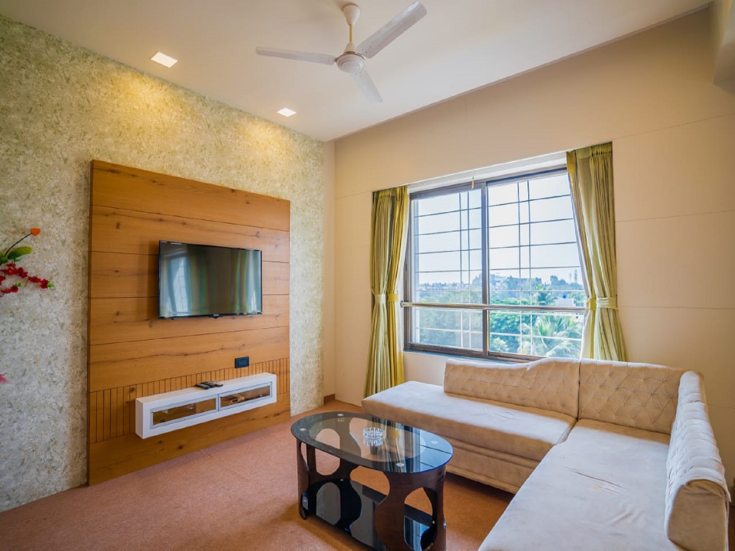 A living room with a wooden wall panel, a flat-screen TV mounted above a built-in shelf, a glass coffee table, and a beige sofa.
