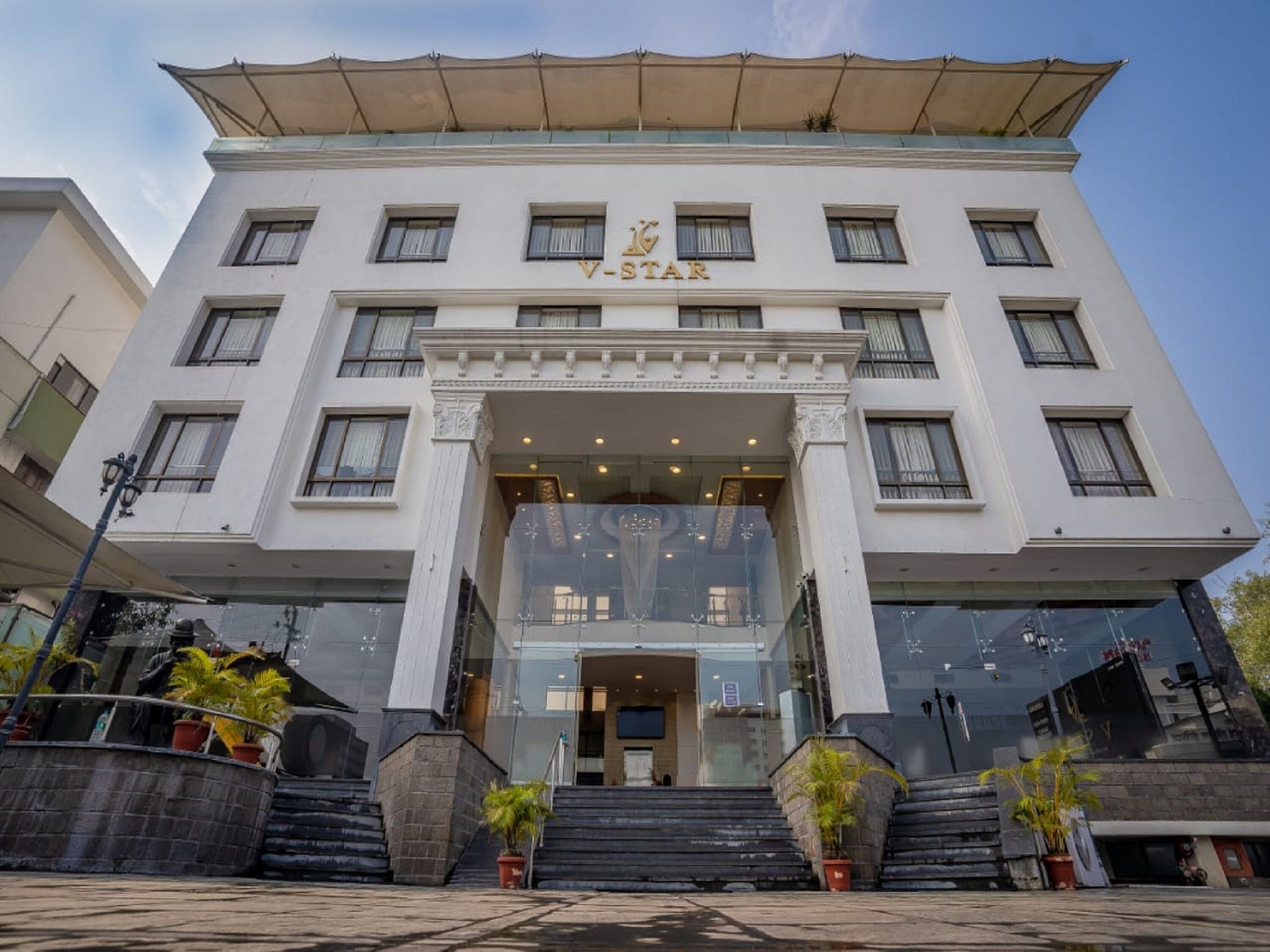 Un hôtel de la chaîne V-Star, doté d’une façade moderne, de plusieurs étages et d’une entrée imposante agrémentée d’un escalier et de plantes en pot.