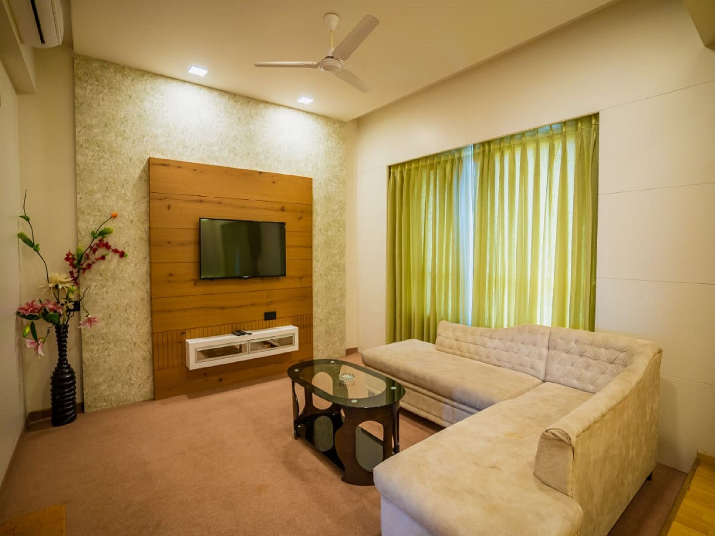 A living room with a sofa, a glass coffee table, a TV mounted on a wooden panel, and a green curtain.