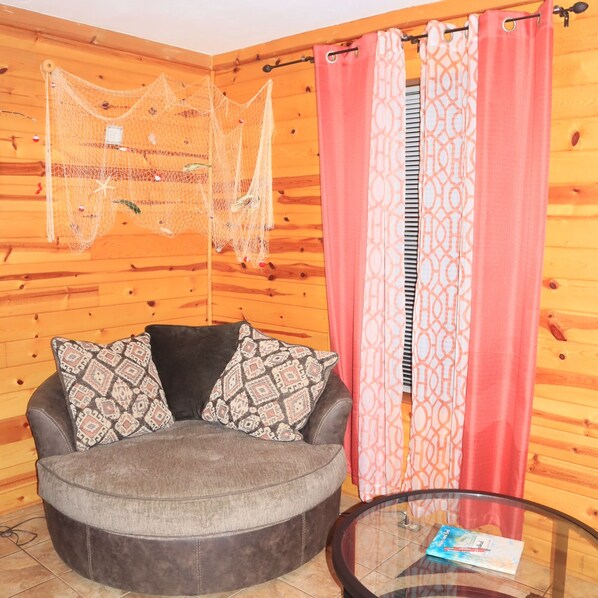A cozy room with a round grey sofa, patterned cushions, a glass coffee table, and a wooden wall with a netted decoration.