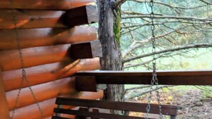 Un banco de madera con estructura metálica, suspendido por cadenas, fijado a una estructura de madera con troncos horizontales.