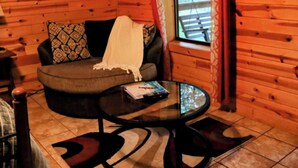 Una habitación acogedora con paredes de madera, un sofá gris con cojines estampados, una mesita de centro de vidrio y una ventana con persianas.