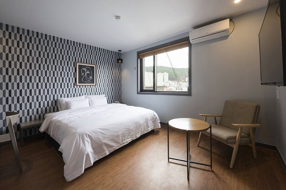 A modern hotel room with a geometric patterned wall, a bed with white linens, a round table, a chair, and a window with a view of buildings.