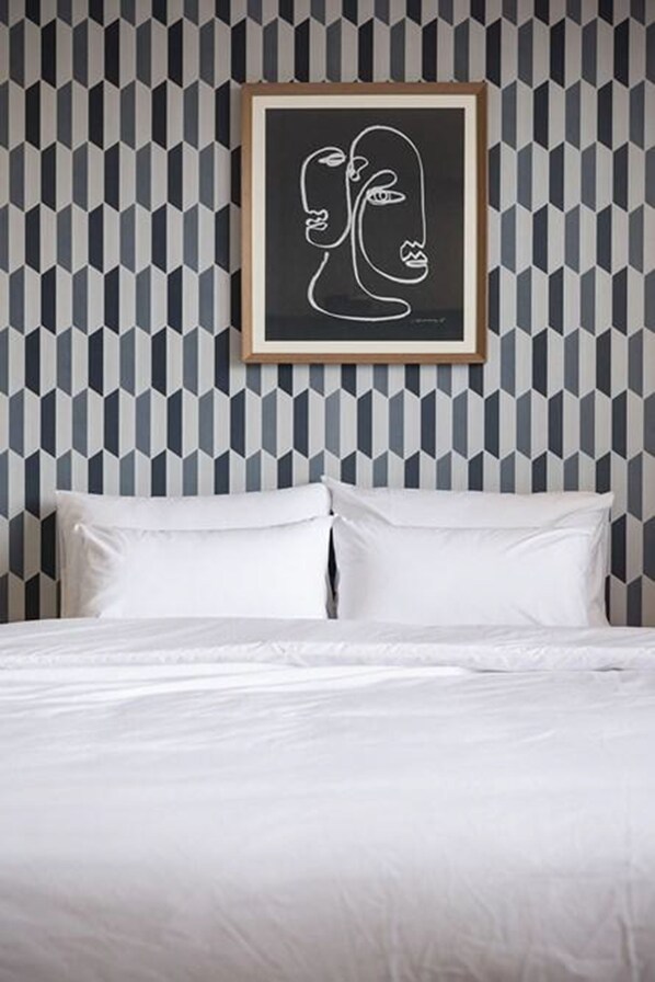 A bed with white bedding and pillows, a geometric patterned wallpaper, and a framed artwork above the bed.