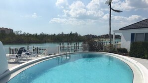 A clear swimming pool with a curved edge, surrounded by lounge chairs and a view of a body of water and buildings in the background.