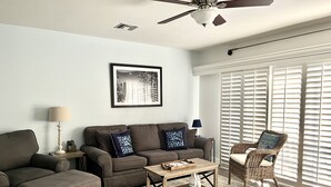 A living room with a sofa, ottoman, coffee table, and a wicker chair. There is a ceiling fan, a framed picture on the wall, and a window with blinds.
