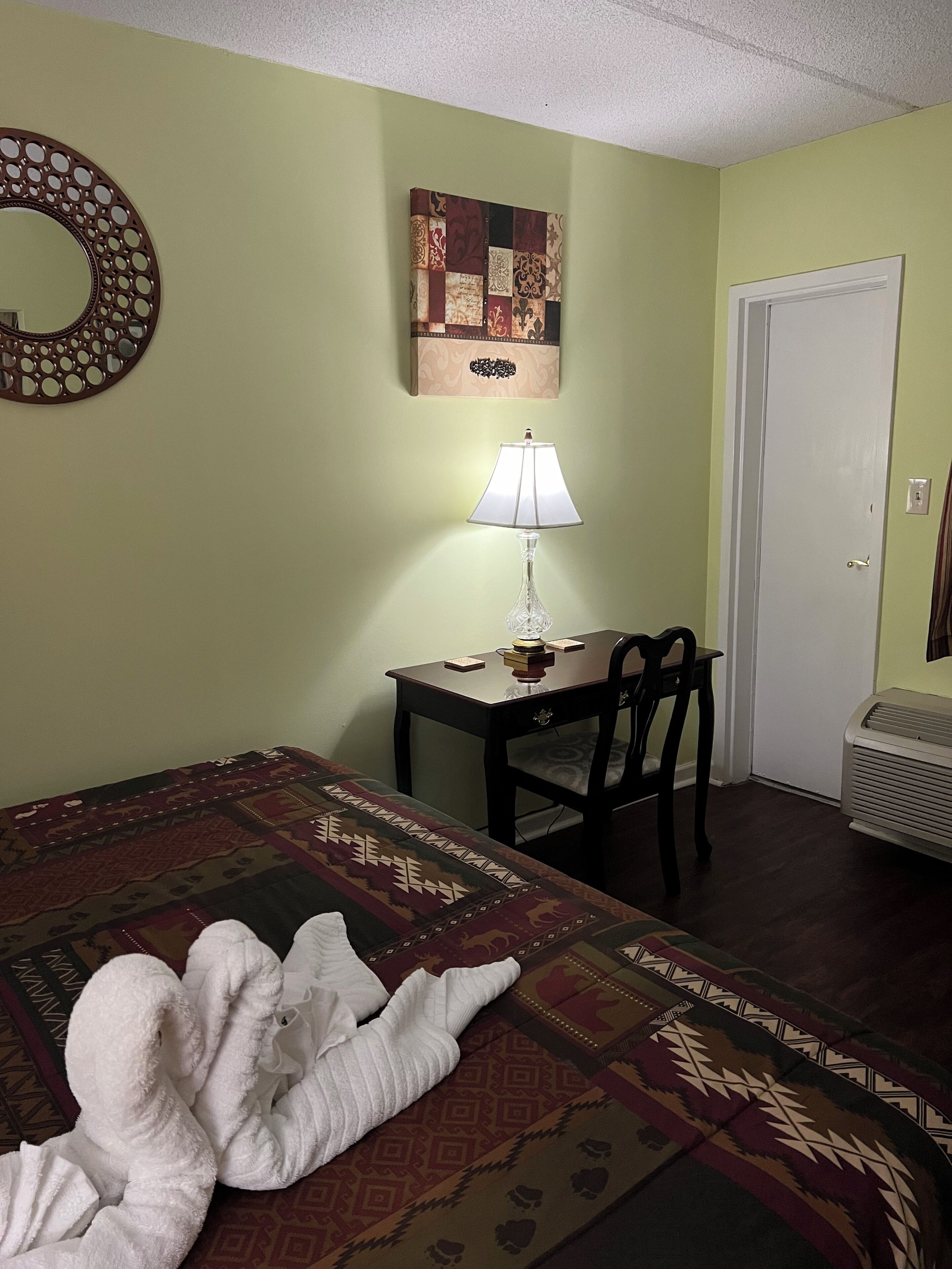 A hotel room with a bed, a table with a lamp, a chair, a wall decoration, and a door.