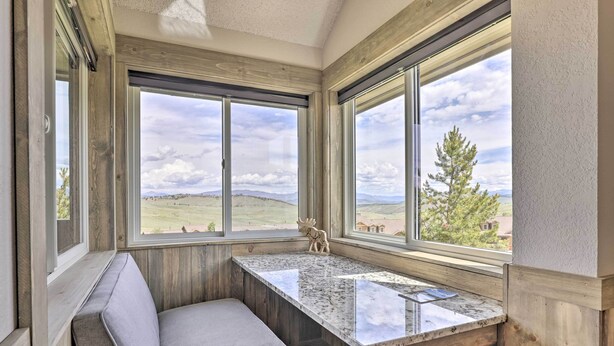 A cozy reading nook with a cushioned bench and a marble-top table, large windows offering a scenic view of a hilly landscape.