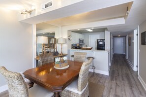 A dining area with a wooden table and wicker chairs, a kitchen in the background, and a hallway leading to a door.