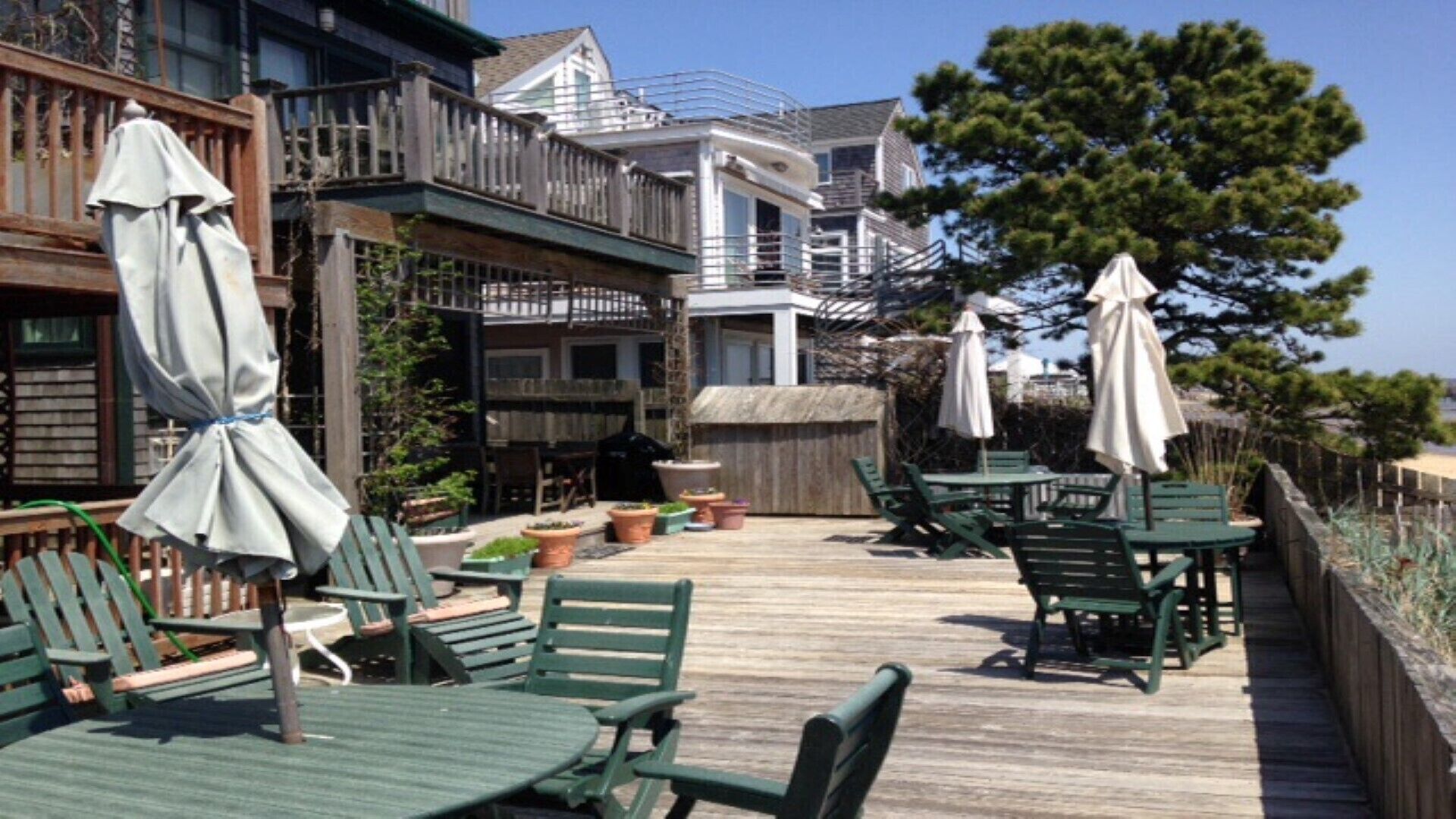 A wooden deck with green chairs and tables, surrounded by potted plants and umbrellas.