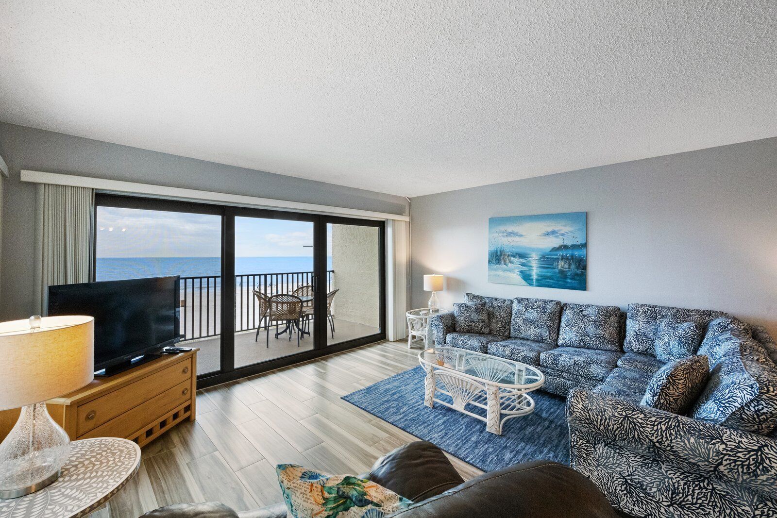 A balcony with a view of the sea, a sofa with patterned cushions, a television on a wooden cabinet, and a lamp on a side table.