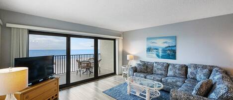 A balcony with a view of the sea, a sofa with patterned cushions, a television on a wooden cabinet, and a lamp on a side table.