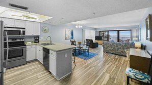 Une cuisine moderne dotée d’un îlot central, un coin repas avec une table et des chaises, un salon avec un canapé et un balcon offrant une vue sur l’océan.
