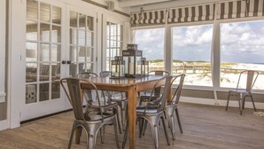 A dining area with a wooden table and metal chairs, large windows with beach views, and a ceiling with exposed beams.