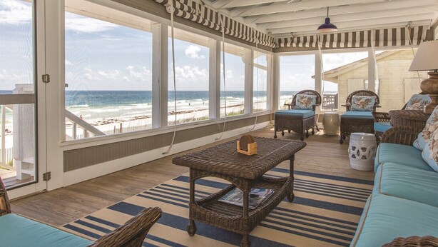 A covered outdoor seating area with wicker furniture, large windows overlooking the beach, and a view of the ocean.