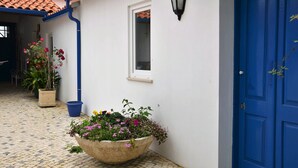 Un edificio con puerta azul, pared blanca, una ventana y una maceta grande con flores.