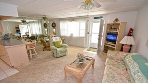 A living room with a sofa, a coffee table, and a television. There is a dining area with chairs and a table. - Pretty, spacious 2-bedroom condo with free WiFi and adorable coastal decor located uptown and only a half a block from the beach! (Ocean City)