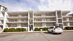 A multi-story building with balconies, a parking lot, and a white car parked in front. - Pretty, spacious 2-bedroom condo with free WiFi and adorable coastal decor located uptown and only a half a block from the beach! (Ocean City)