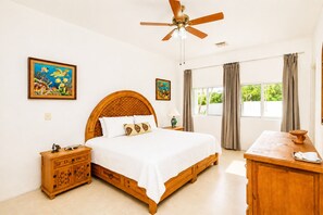 Une chambre à coucher avec un lit en bois, une table de chevet, un bureau, un ventilateur de plafond et deux fenêtres avec des rideaux.