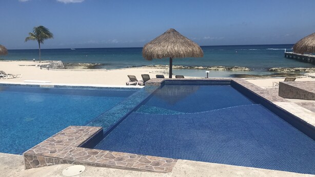 Ein Pool mit Steinrand, Liegestühlen, einem Reetdachschirm und Blick auf den Strand.