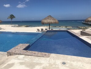 Ein Pool mit Steinrand, Liegestühlen, einem Reetdachschirm und Blick auf den Strand.