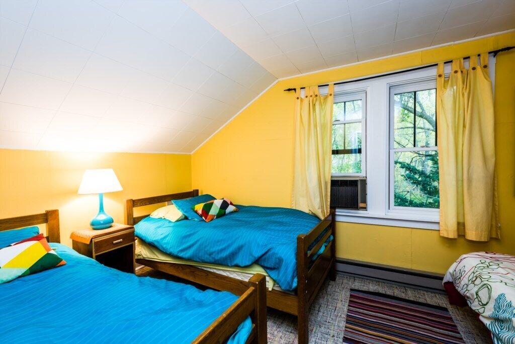 A room with two beds, a window with curtains, a lamp, and a view of greenery outside.