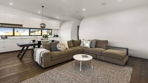 A modern living room with a sectional sofa, a dining table, and a kitchen area in the background. - Yellow Door Coburg | Boutique Accommodation | Sleeps up to 16 guests (Coburg)