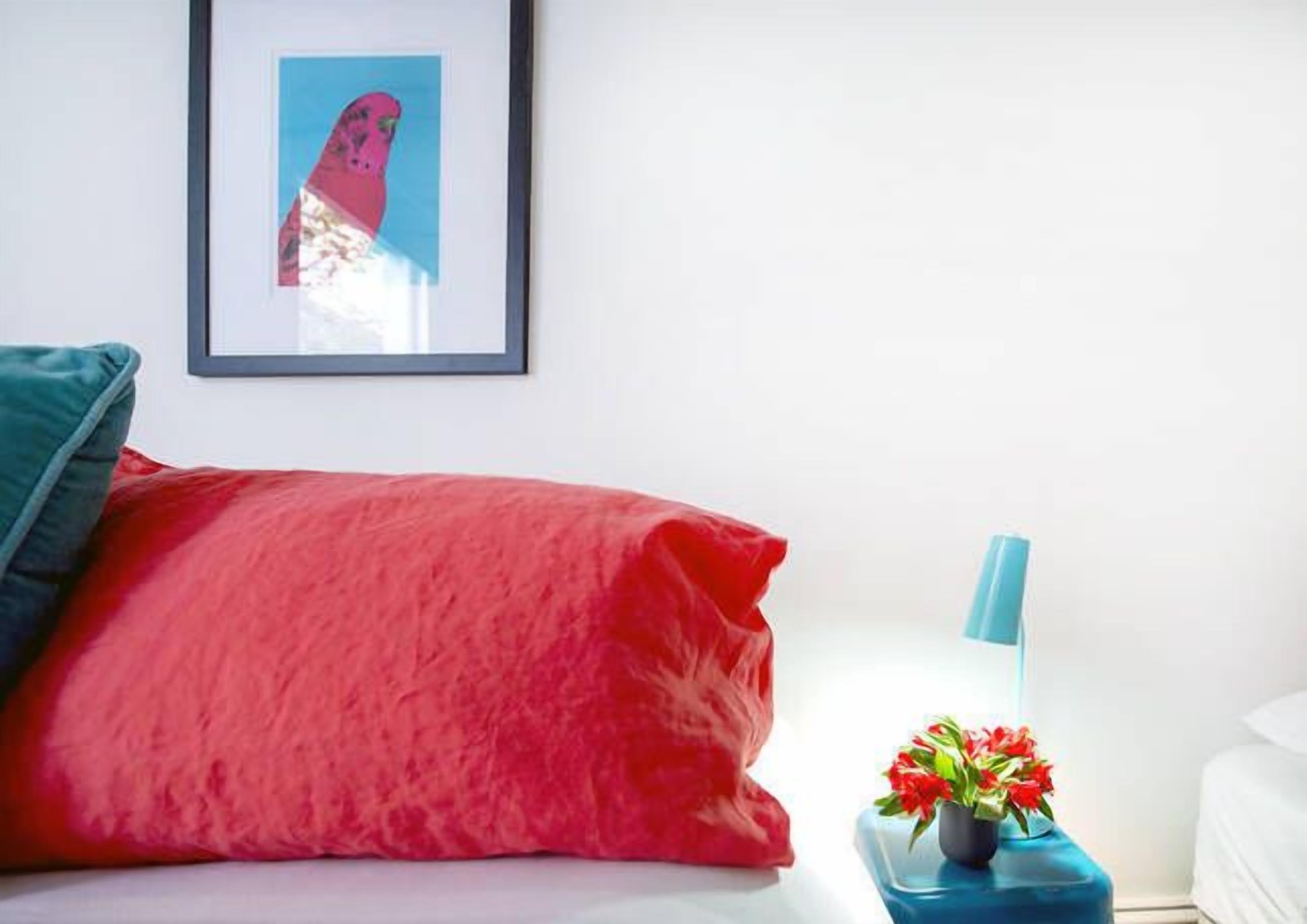 Ein Schlafzimmer mit roter Bettdecke, einem gerahmten Kunstwerk an der Wand, einem blauen Nachttischchen mit einer roten Blumenvase und einem weißen Bett.