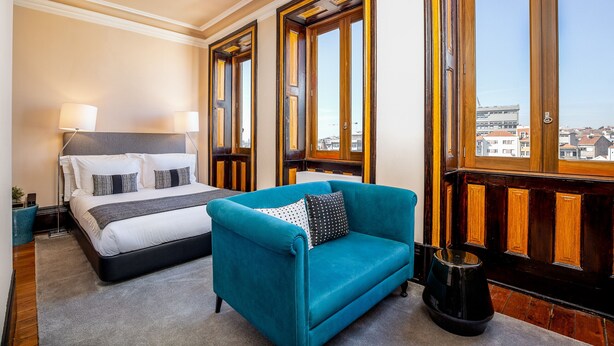 A hotel room with a bed, a teal armchair, a wooden door, and a view of buildings. - Original Studio in Bolhão Market (Oporto)