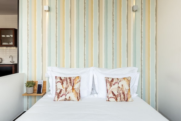 A bedroom with a striped wall, a bed with white bedding, two pillows with a leaf pattern, a bedside table with a plant and a digital clock, and a small shelf with dishes.