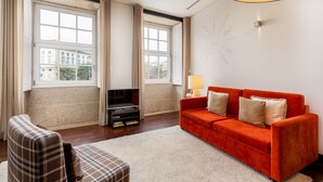 Un salon avec un canapé rouge, un fauteuil à motifs, une télévision et une grande fenêtre avec des rideaux.