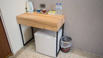 A compact kitchenette with a small fridge, a kettle, two cups, a plate, and two water bottles on a wooden table.