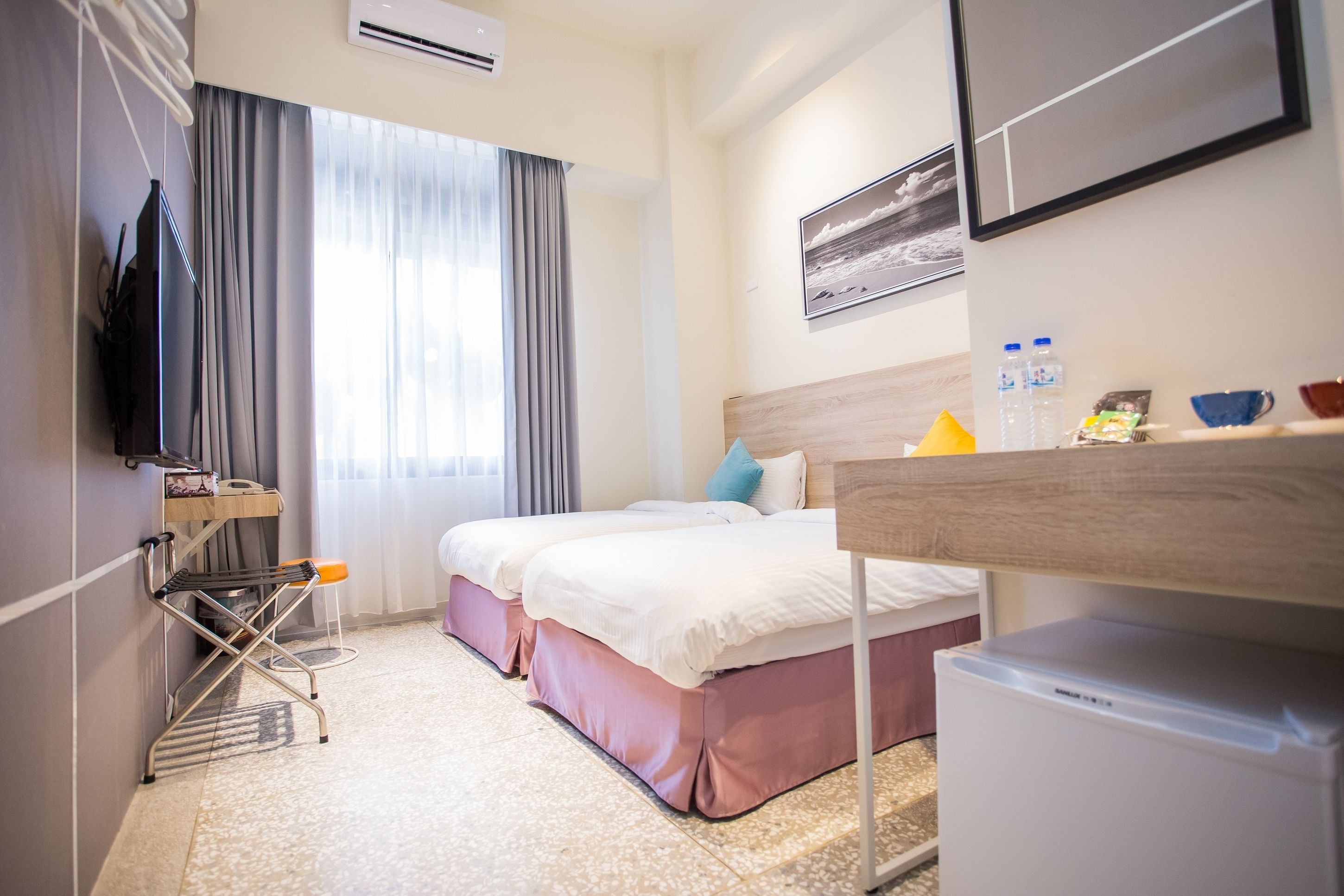 A hotel room with two beds, a TV, a mini-fridge, a desk, and a window with curtains.