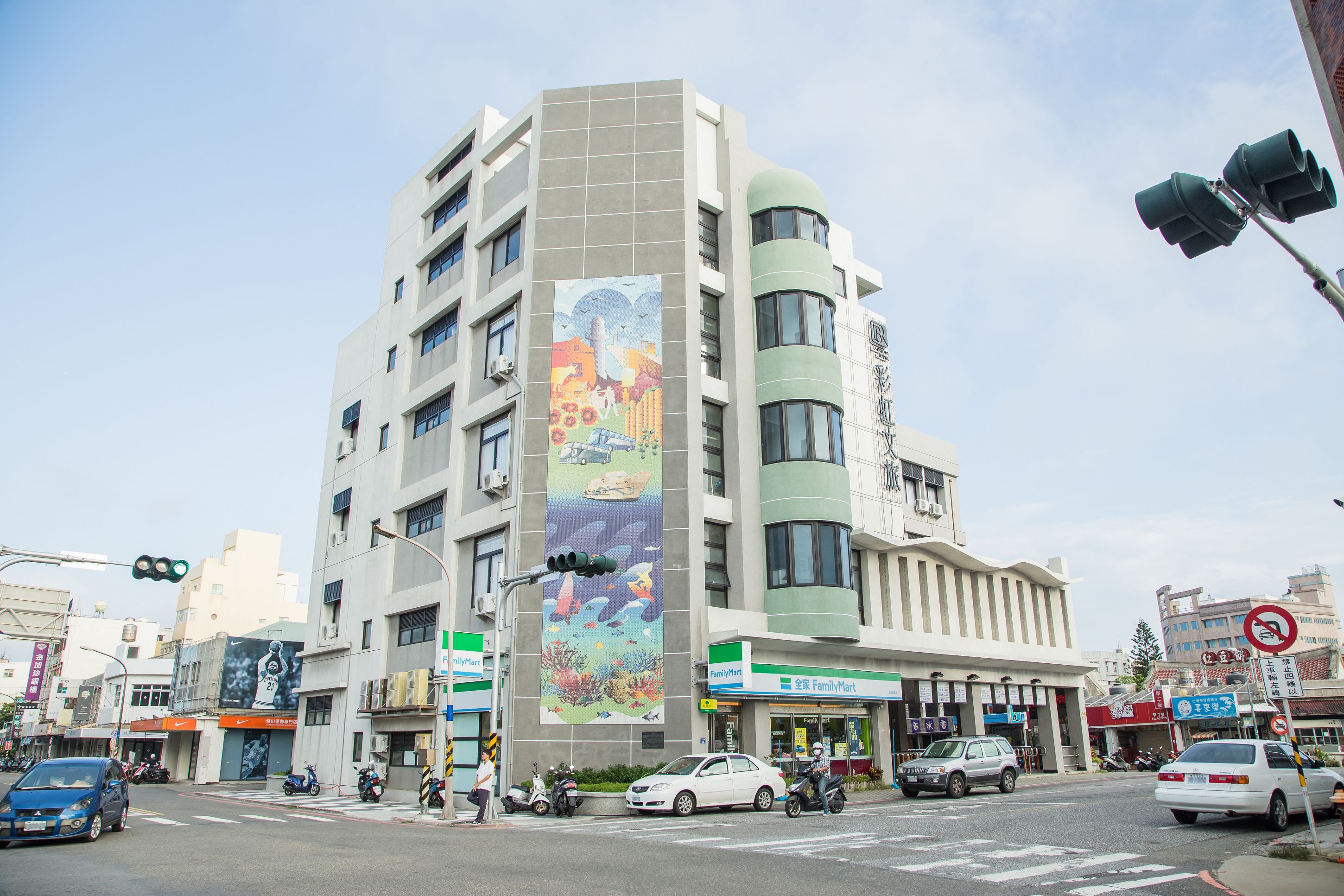 A multi-story building with a large mural on the side, a FamilyMart store, and various vehicles at the intersection.